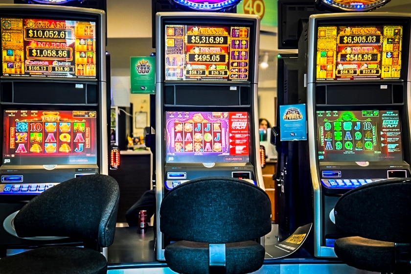 Three poker machines in a club with brightly coloured screens showing different gambling games, with three empty stools in front