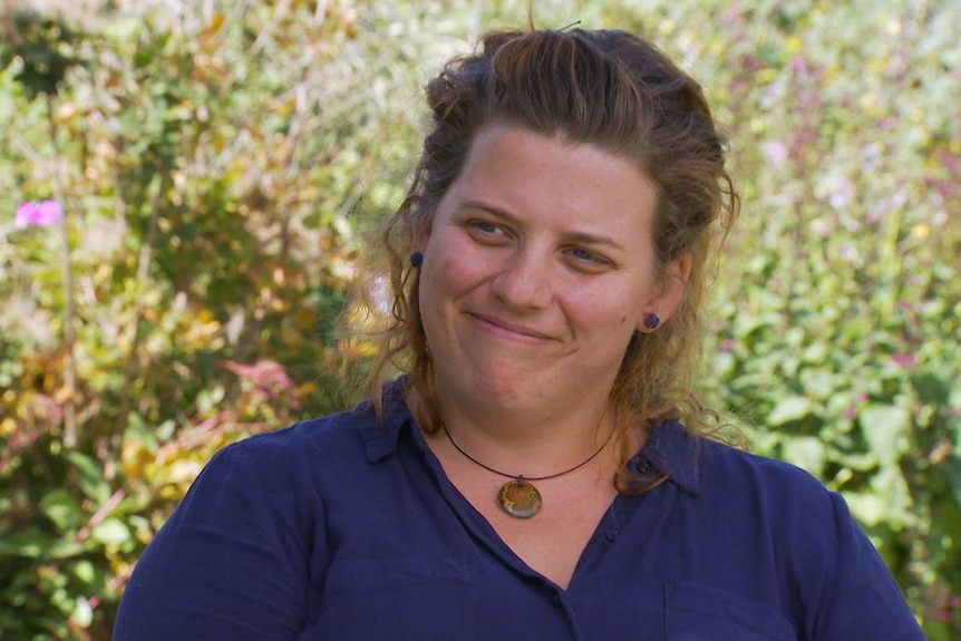 A woman smiles as she sits outside in shade. The background is a garden in sunshine.