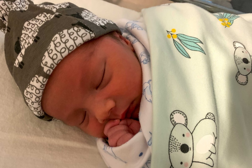 A baby sleeps while holding his fist to his mouth and wrapped in a blanket.