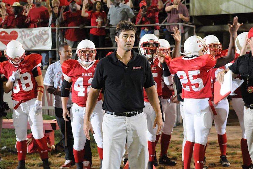 A US football coach stands in front of players and a cheering crowd.