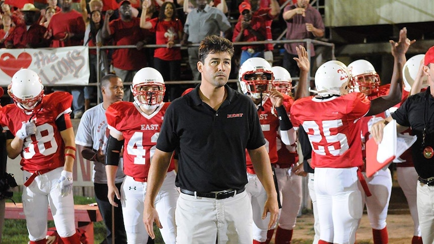 A US football coach stands in front of players and a cheering crowd.