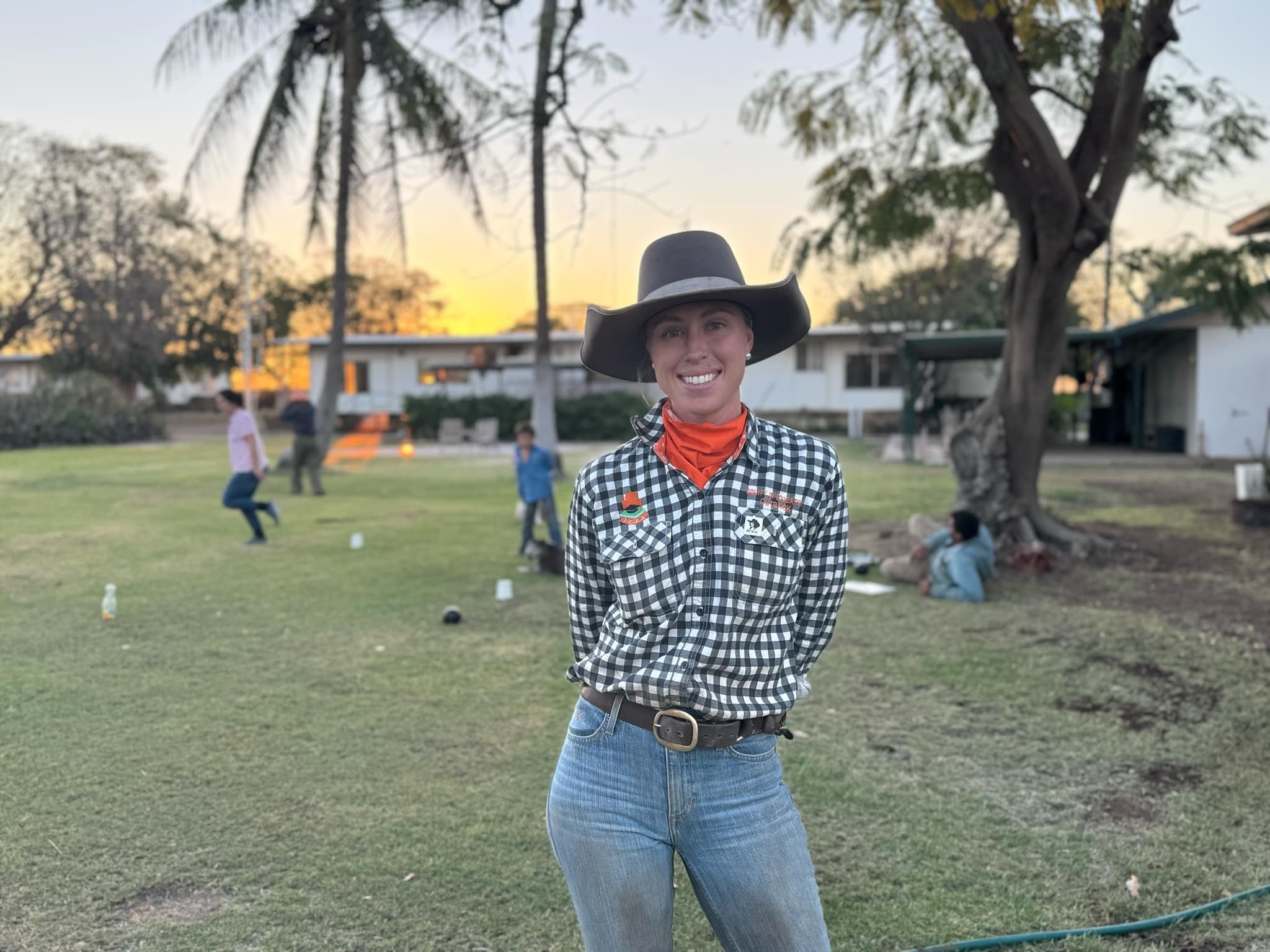 Woman standing on grassy lawn at sunset with children playing in background.