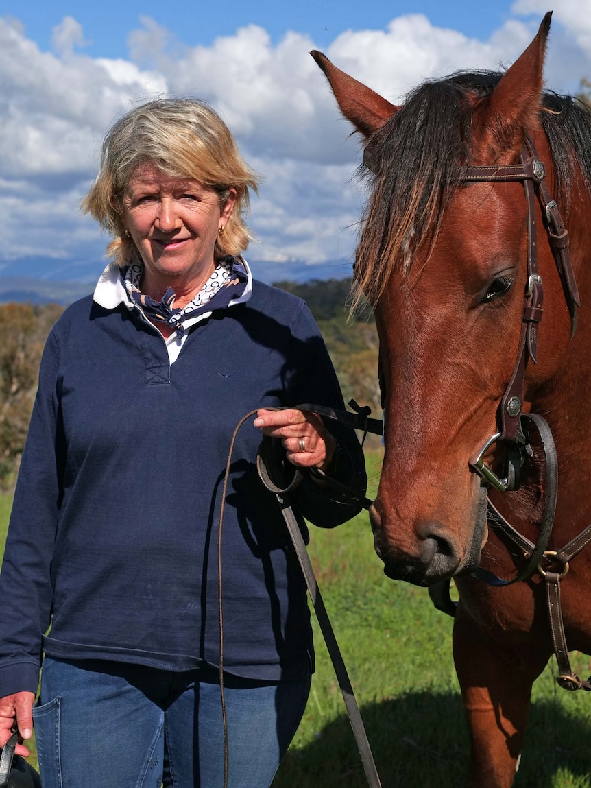 A woman smiles as she stands next to a brown horse, holding its reins.