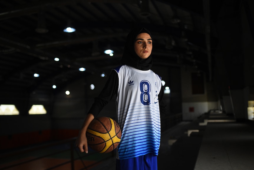 A young woman wearing a head scarf holds a basketball and looks out of the stadium. 