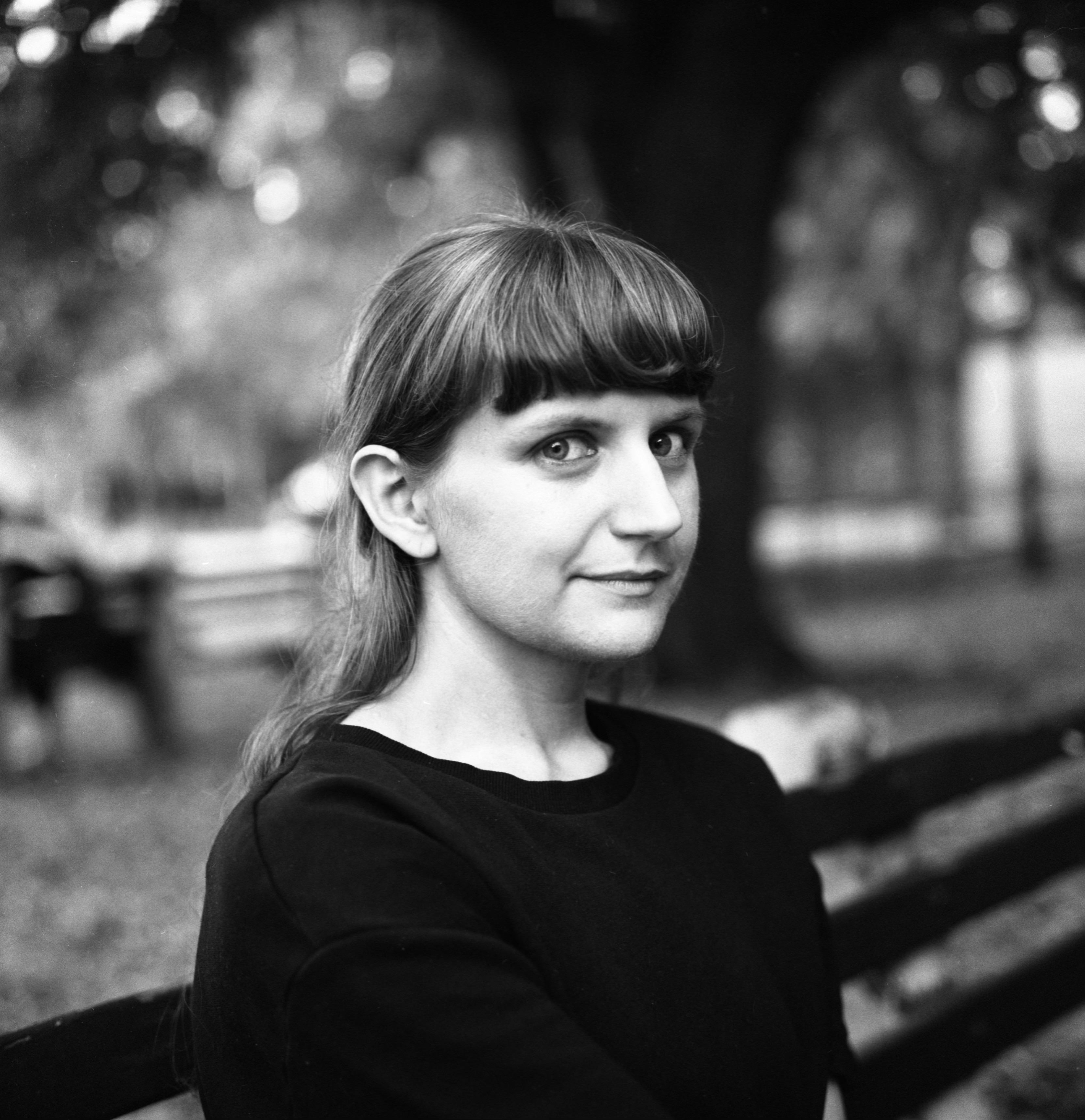 A black and white headshot of a woman outside. She looks side on towards the camera, has a fringe and long hair behind her ears.