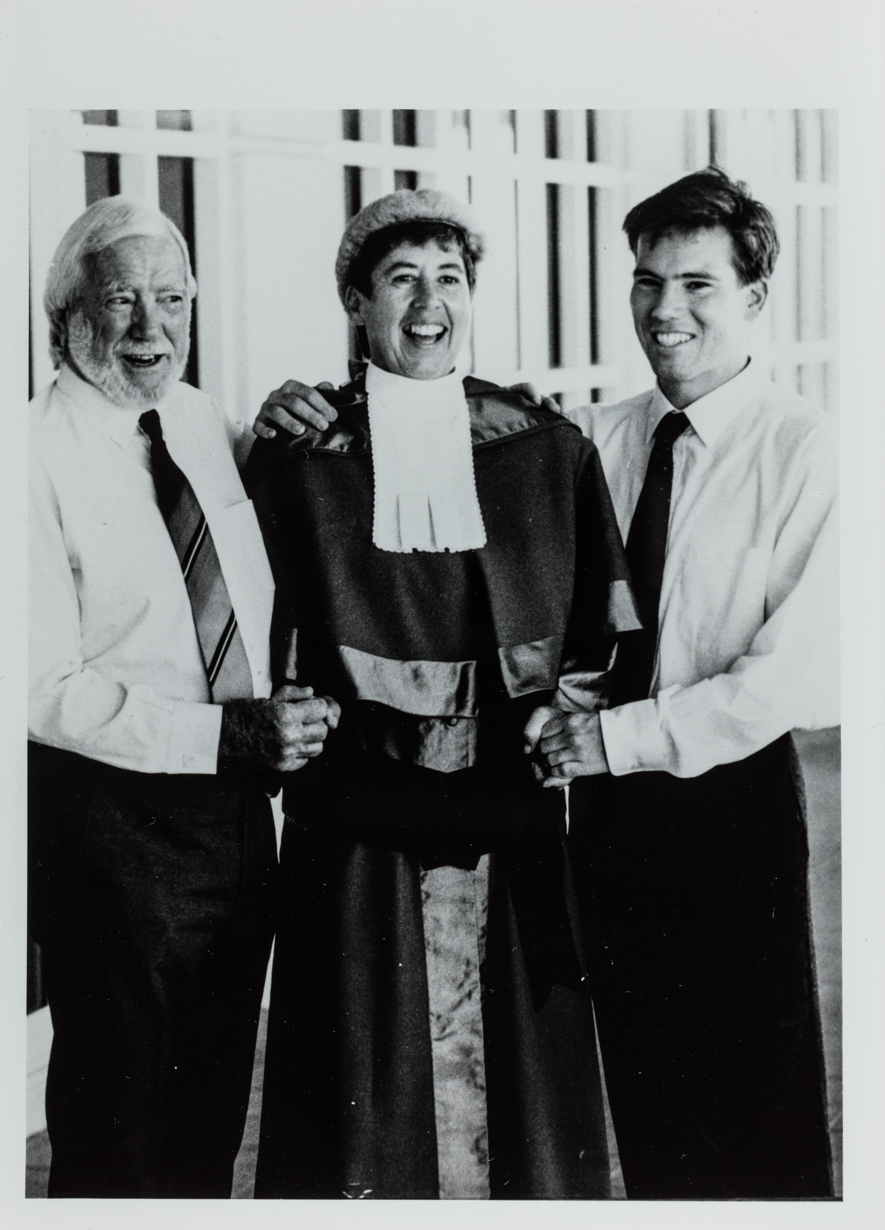 A black and white photo of a woman wearing a judge's robe and wig standing between two men
