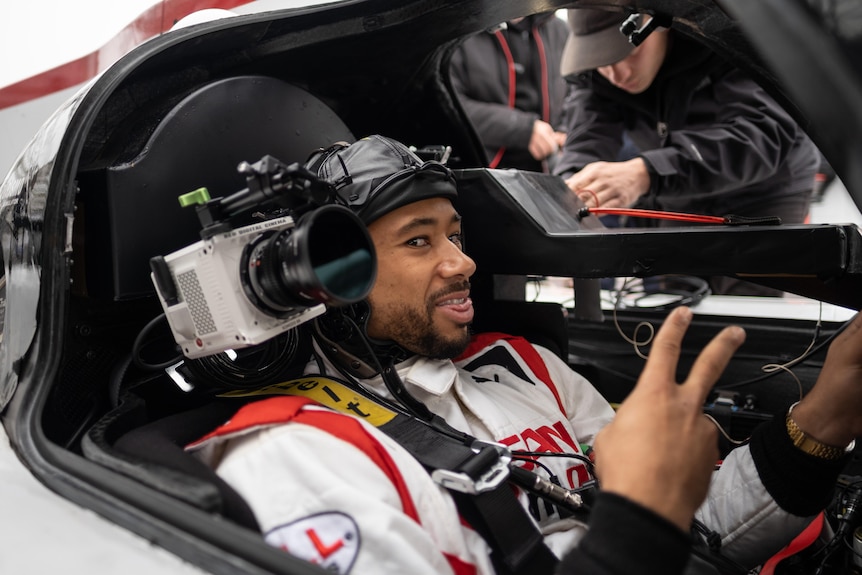 Actor Archie Madekwe smiling with his tongue out in a race car cockpit with a camera mounted by his shoulder.