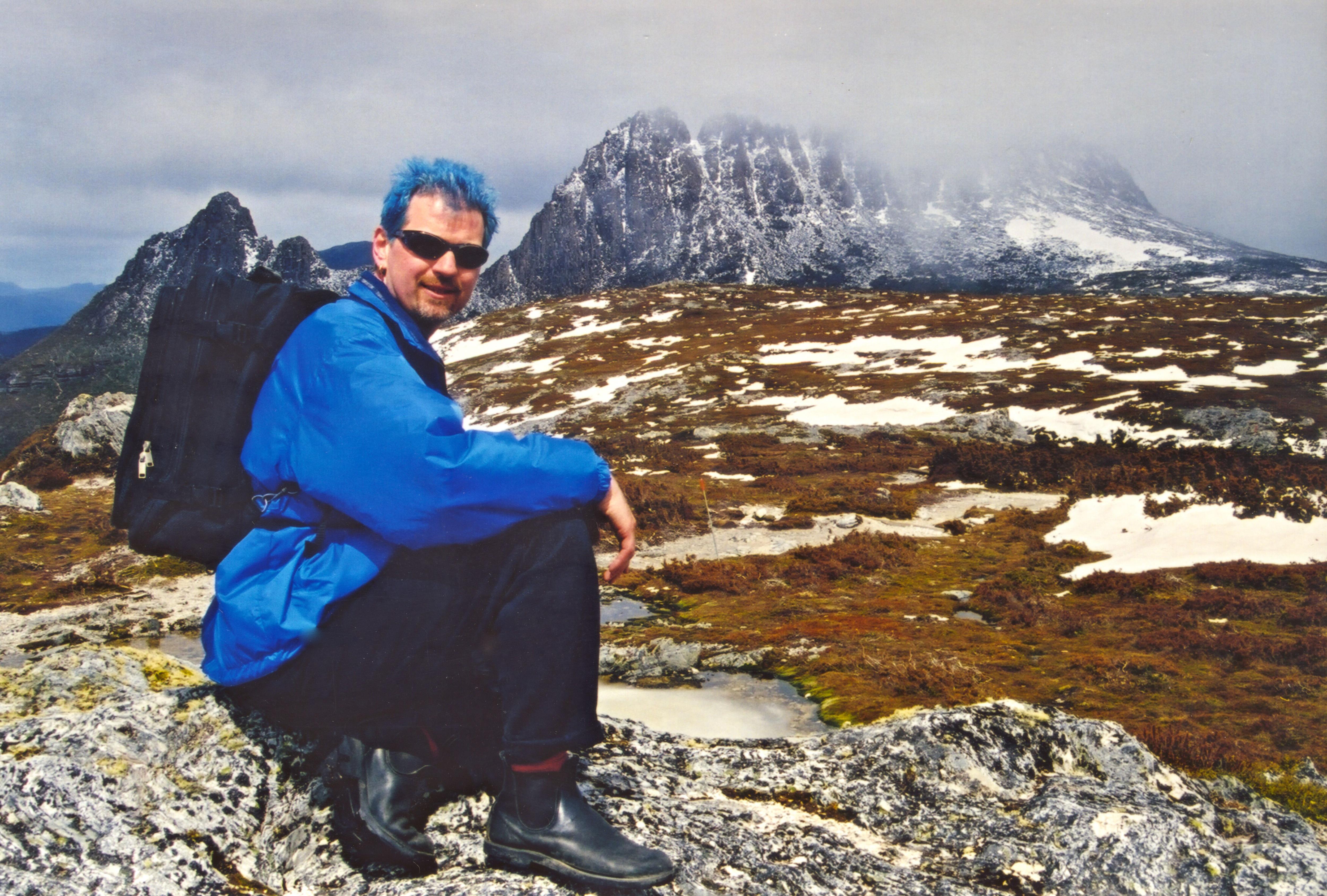 Vintage photo of man with bright blue hair squatting down in front of an epic snowcapped mountain