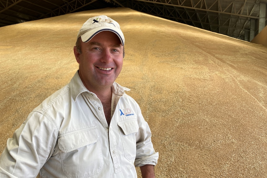 A man in a clay shirt and hat stands in front of a mountain of grain. 