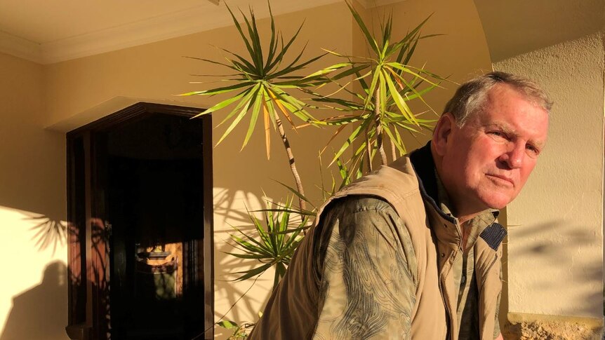 A man leans forward in front of a tall, potted green plant by a front door.