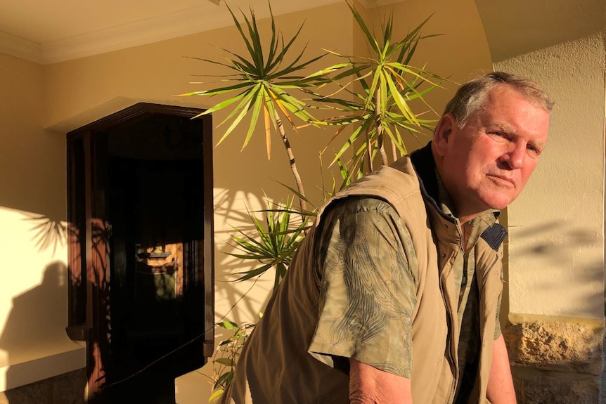 A man leans forward in front of a tall, potted green plant by a front door.