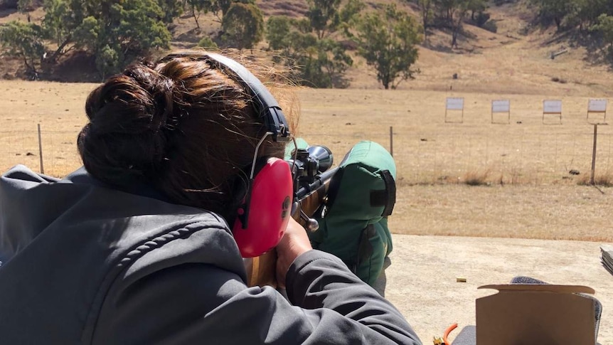 A woman wearing earmuffs puts a gun at a target in a dry paddock.