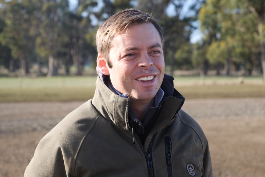 A man in a brown windbreaker stands on a driveway, a line of gum-trees stands in the distance. 