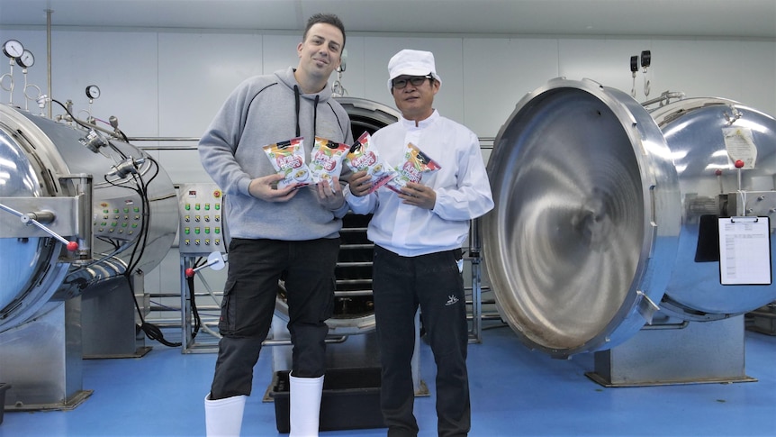 Two men standing in a factory holding packets of apple chips 