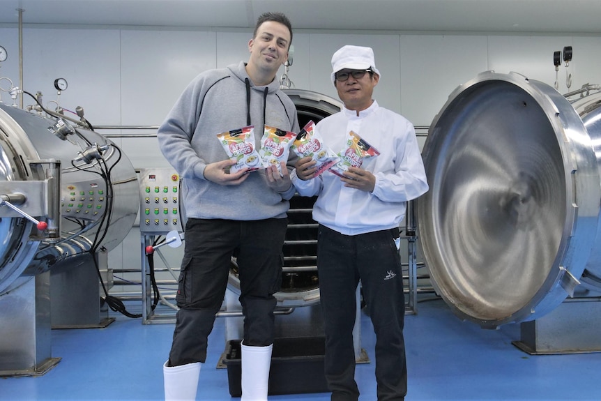Two men standing in a factory holding packets of apple chips 