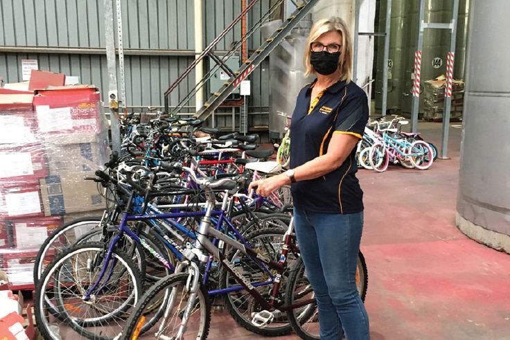 A lady with blond hair and wearing a mask stands next to a bicycle.