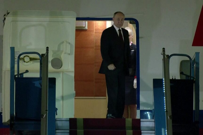 Vladimit Putin stand at an open airplane door at the top of stairs.