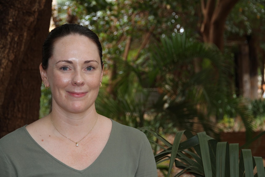 A woman wearing a khaki green dress smiles closed mouth at the camera, with green plants behind her