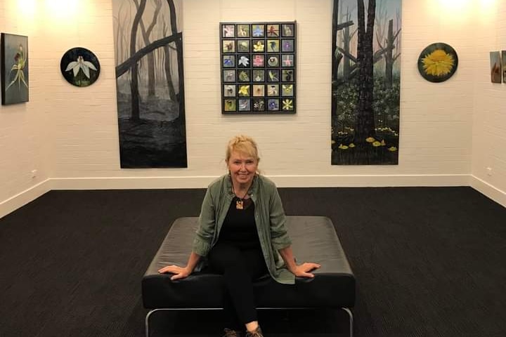 A blonde woman sits on a seat at a gallery, surrounded by paintings of flowers