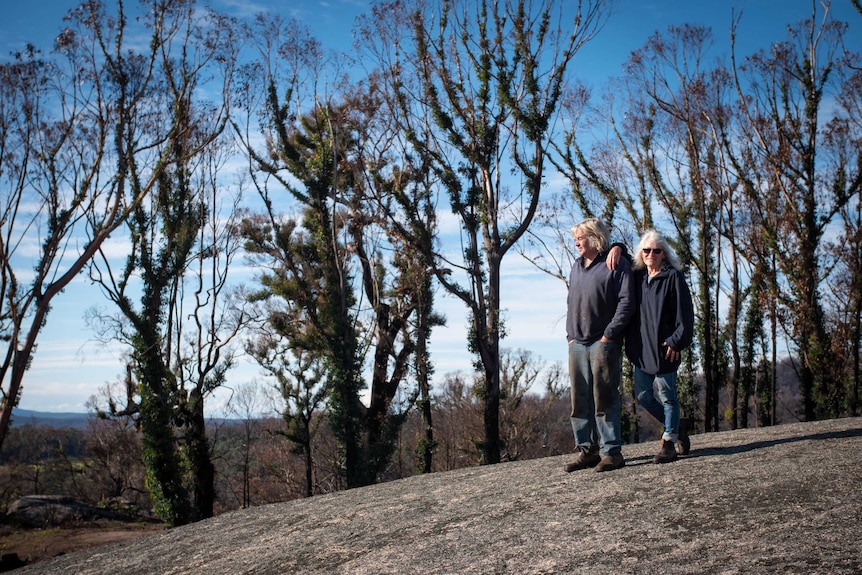 A couple stand on a rock, both with white hair, blue jeans and a blue jumper, behind them are burnt gum trees with re-growth.