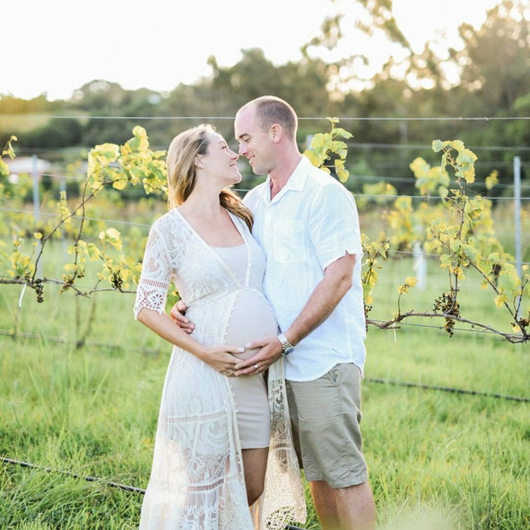 A mand and a pregnant woman embrace among grape vines in a vineyard.
