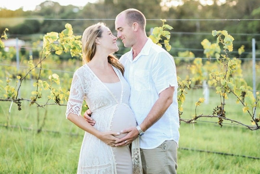 A mand and a pregnant woman embrace among grape vines in a vineyard.