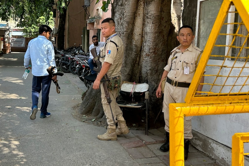 Three armed men, two in uniform, stand next to a large tree next to a building, behind a metal fence.