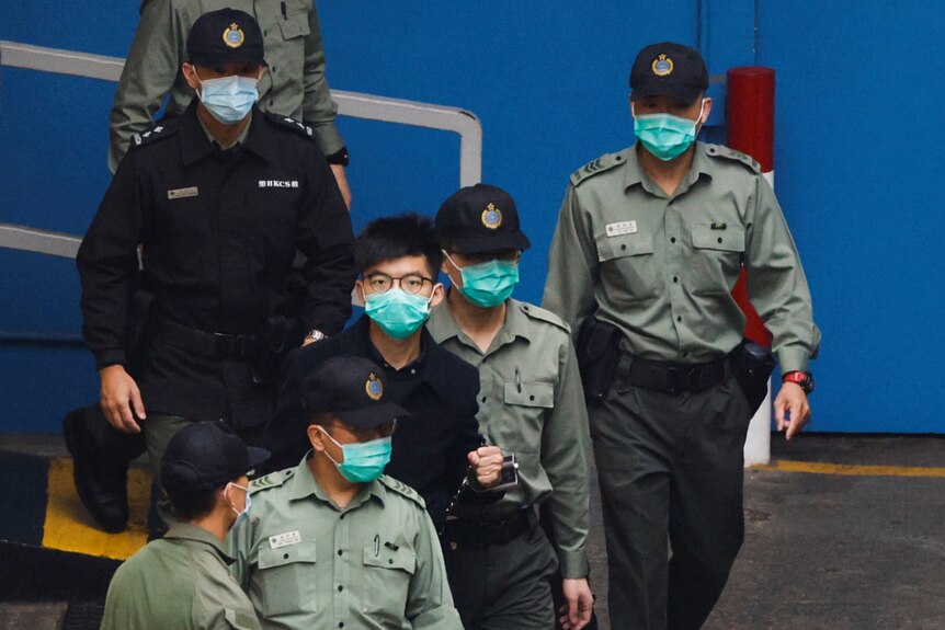 A man wearing glasses and a mask is escorted to a prison van by law enforcement officials.