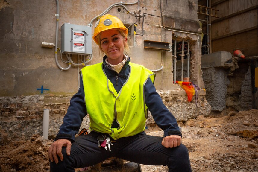 A young woman sitting down at a a building worksite. She's wearing a hard hat