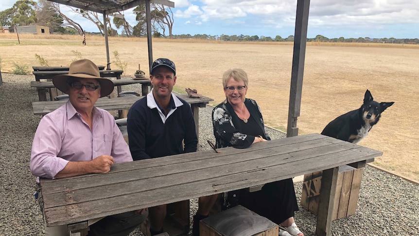 Three people sit at a table outdoors with a working dog.