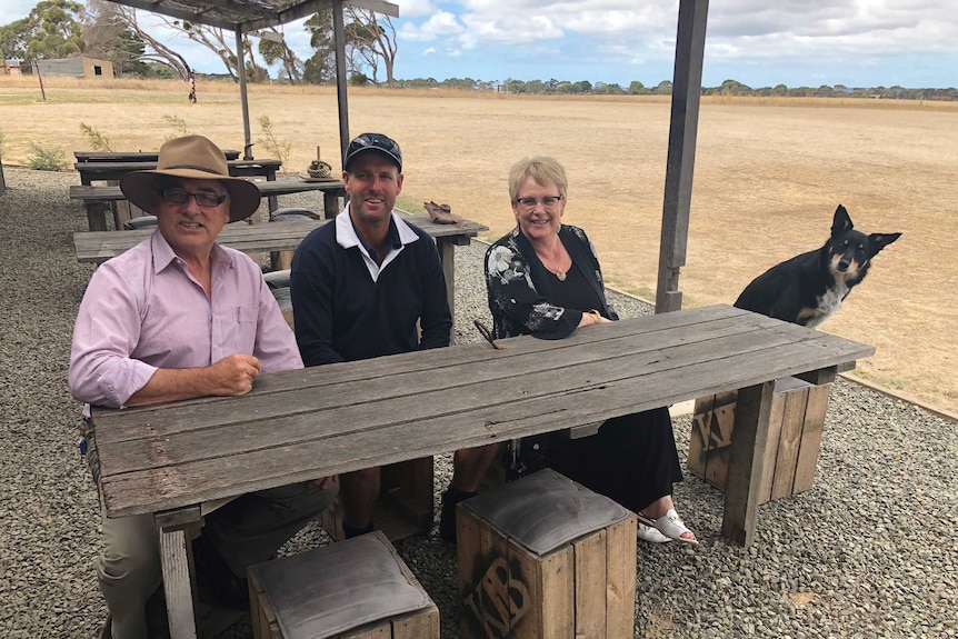 Three people sit at a table outdoors with a working dog.