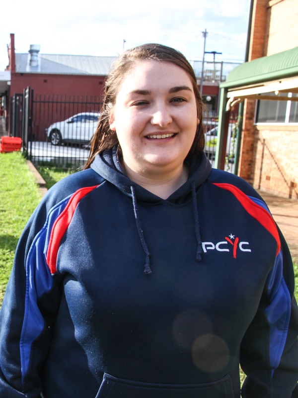 A young woman with straight black hair and a navy top stands outside a PCYC building, smiling