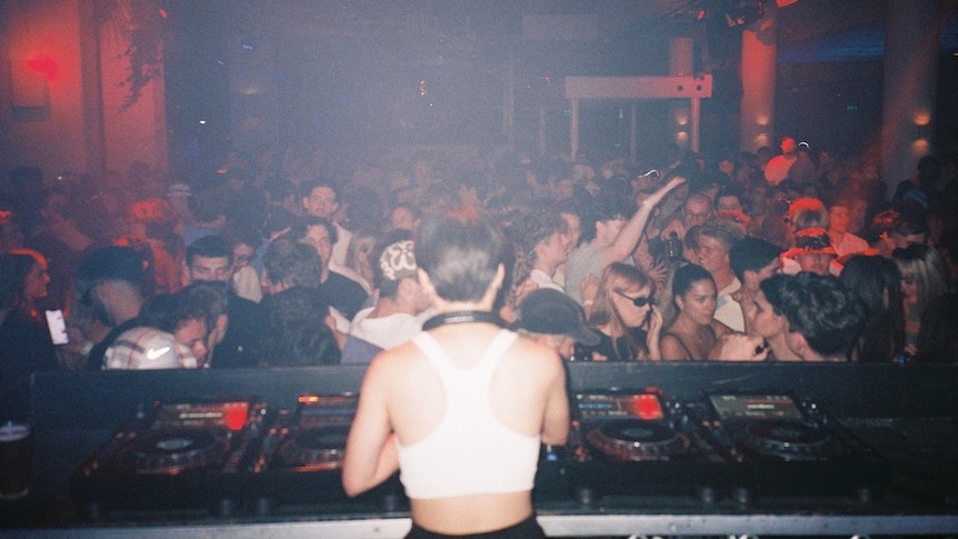A femme presenting DJ wearing a white singlet is standing behind some DJ decks facing a crowd full of dancing people in a club. 