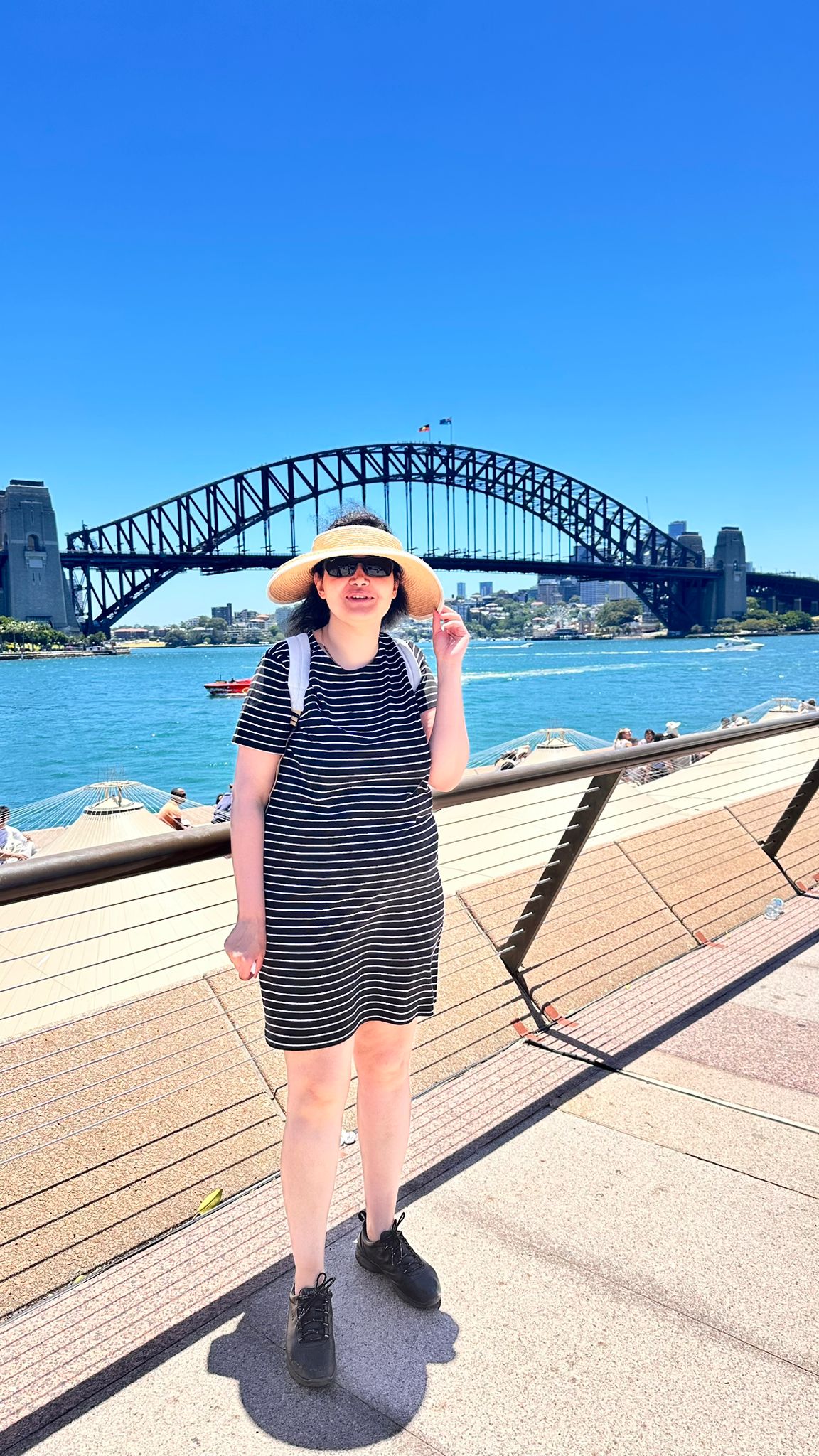 Uma mulher de óculos escuros e chapéu fica de costas para a Sydney Harbour Bridge em um dia ensolarado.