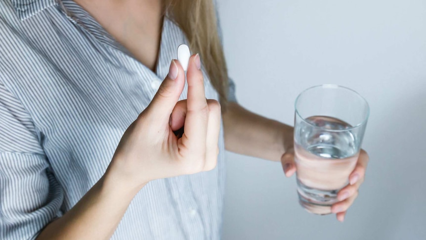 A woman holds a pill in one hand and a glass of water in the other.