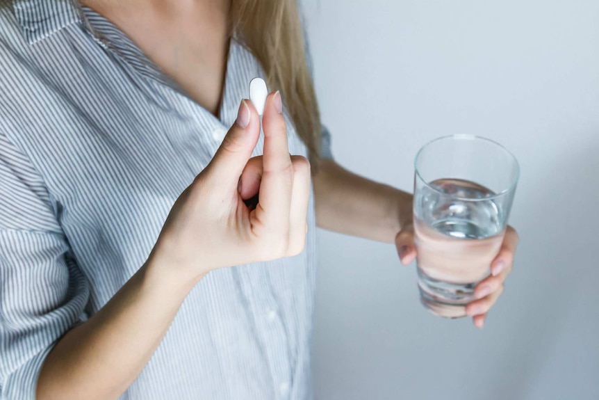 A woman holds a pill in one hand and a glass of water in the other.