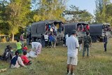 People stand and sit around on grass after getting out of army trucks. 