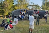 People stand and sit around on grass after getting out of army trucks. 