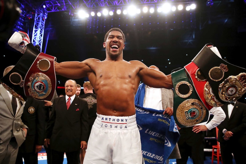 Anthony Joshua stands with his arms outstretched with five boxing belts draped over his arms.