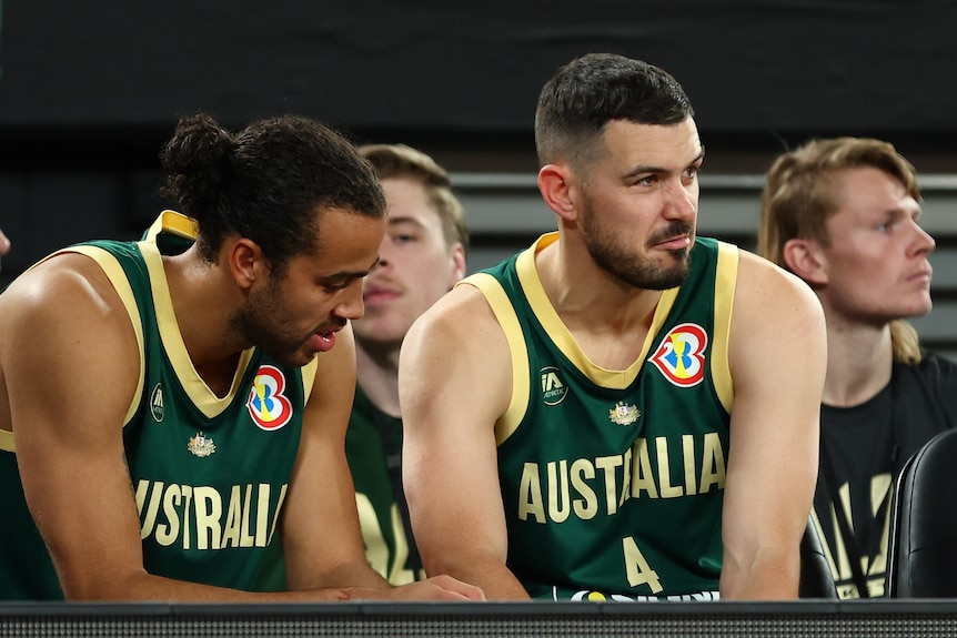 An injured Australian basketballer (right) sits with a teammate on the sidelines watching a game.