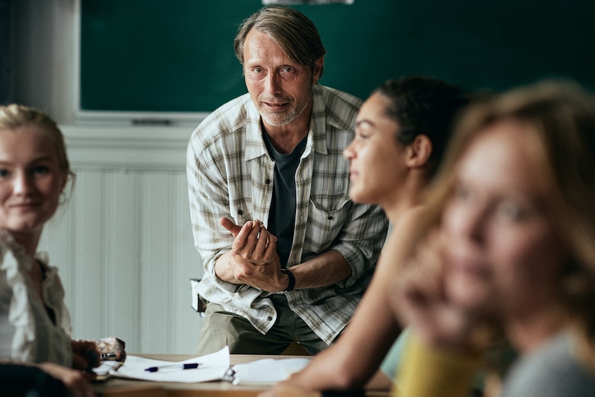 Mads Mikkelsen in checked flannel shirt in classroom, with some female students in front of him, as he talks.