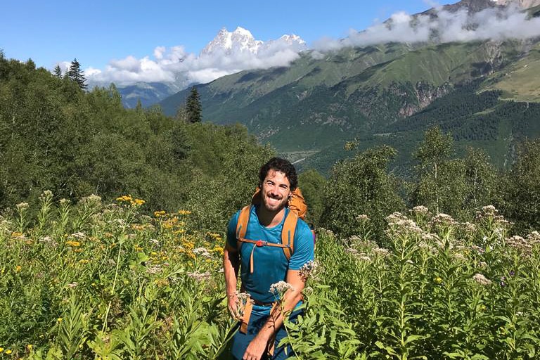 A man with black hair and a beard wearing a backpack smiles on a sunny day, mountains behind him.