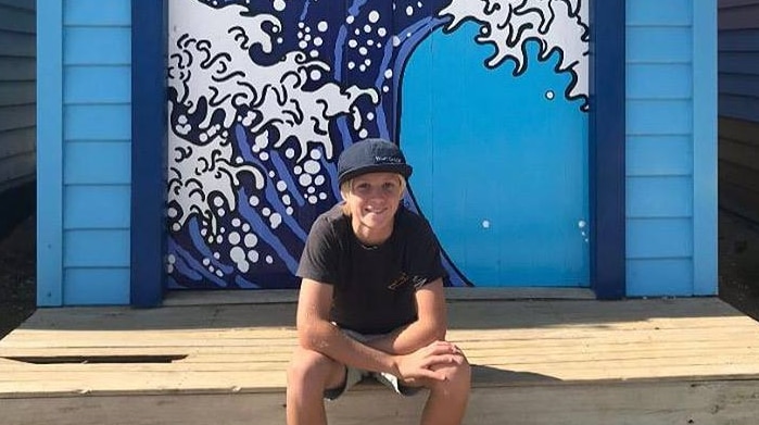 12-year-old Noah Pronk smiles as he sits in front of a small blue beach shack with a wave painted on it.