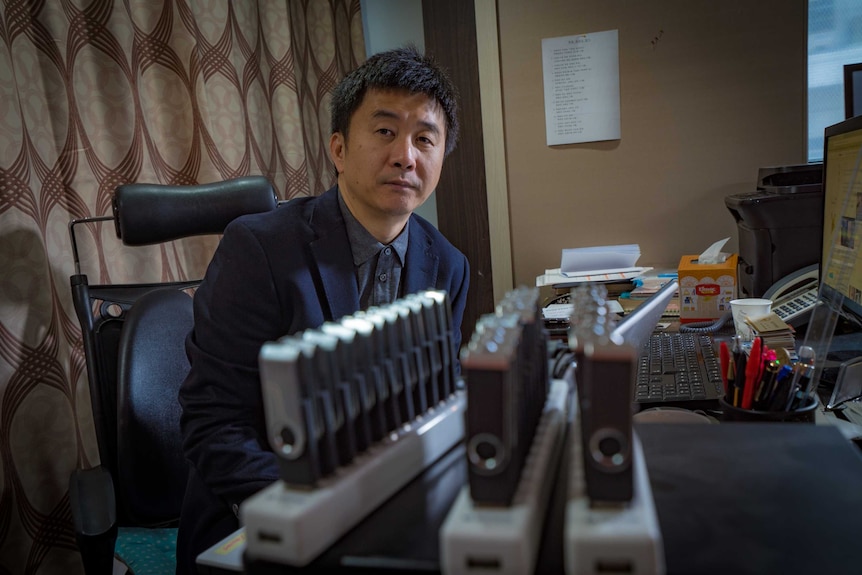 Chol Hwan Kang sits at a desk behind USB hubs used to clone USB sticks.