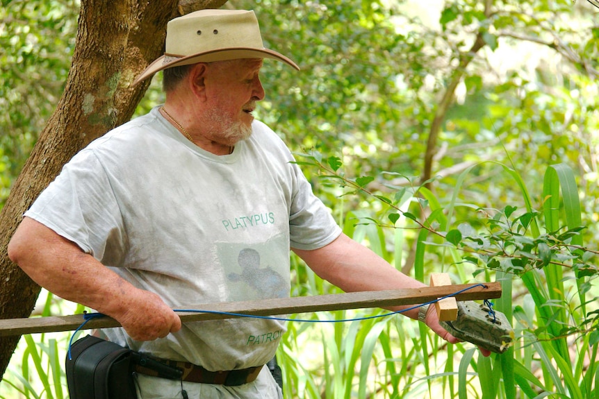 A man holds a wooden stake in one hand while opening the casing of a camera trap with his other.