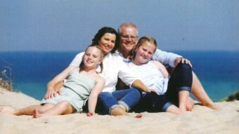 A family of two girls and parents sit on the beach smiling