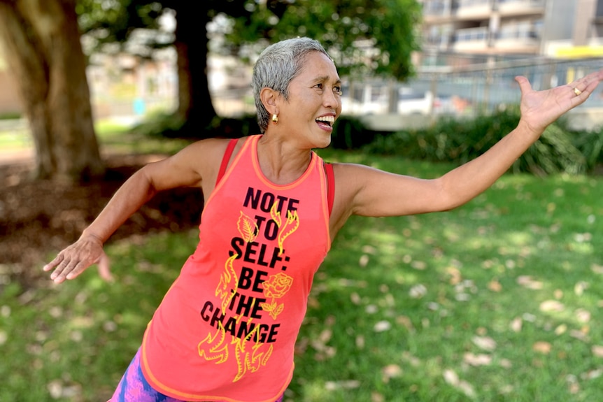 An older, Asian woman wearing a singlet that says "Note to self: Be the change" as she appears to be doing tai-chi