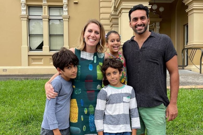 a family with a woman, a man and three young children standing outside of a building and smiling