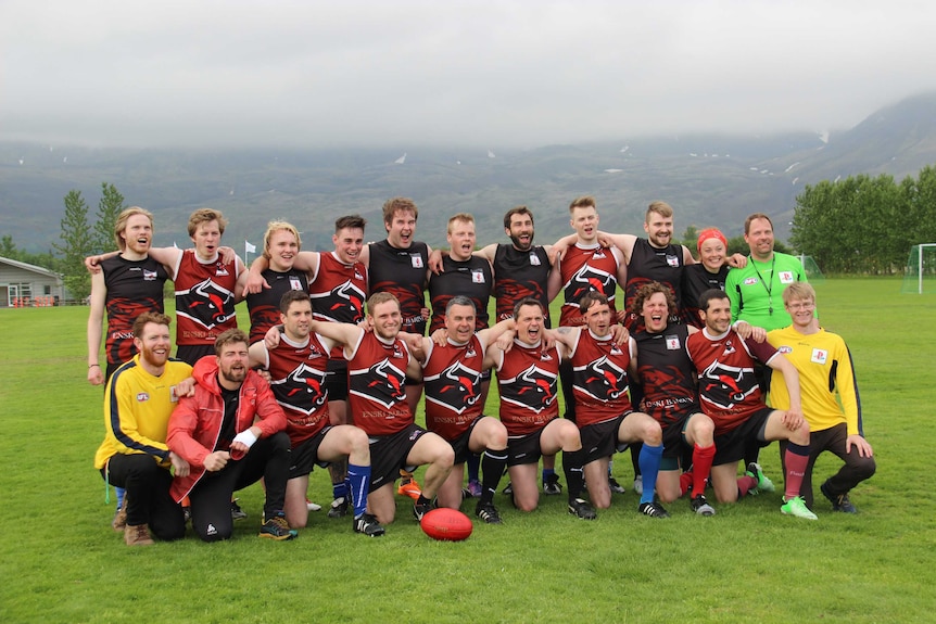 A team of Aussie Rules footballers wearing black and red jumpers pose for a team photo on an oval in Reykjavik, Iceland.