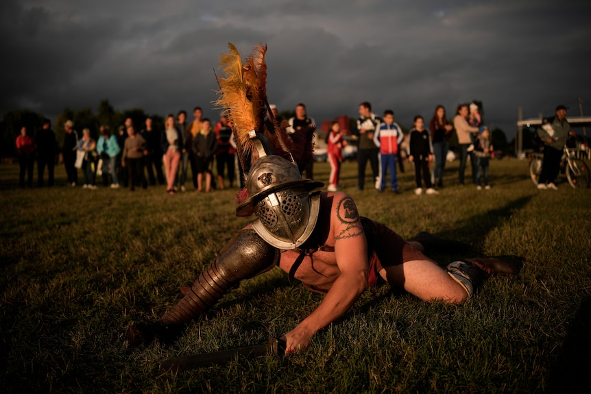A Roman gladiator re-enactor with a helmet on looks up from the ground with members of the public watching on